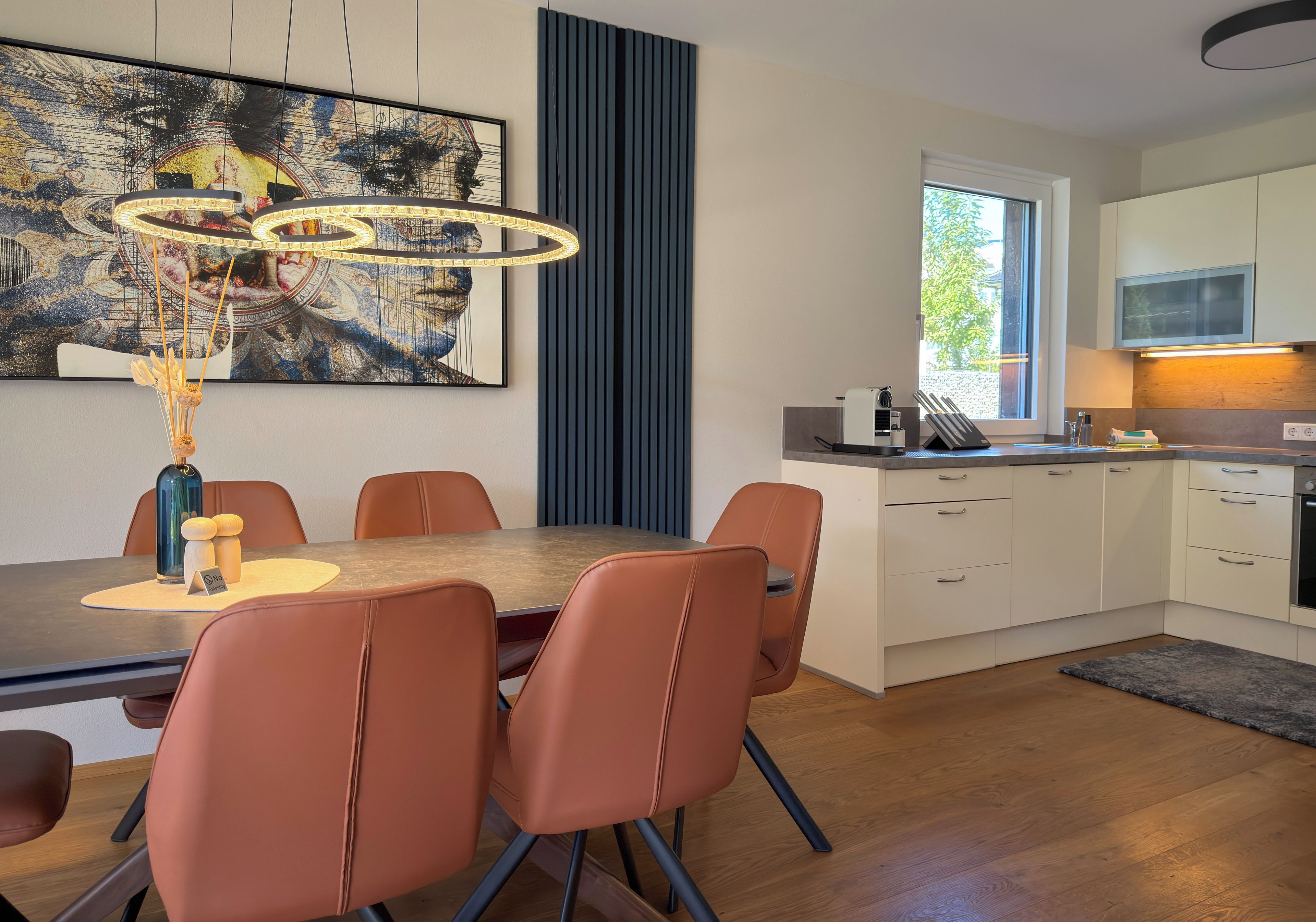 A modern dining area with a table and chairs, a painting on the wall, and a kitchen in the background.