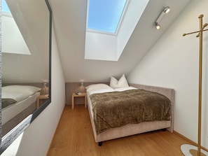 Ein Schlafzimmer mit Dachfenster, einem Bett mit brauner Decke, einem Nachttisch mit Lampe und einer Stehlampe.