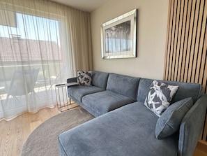 Ein modernes Wohnzimmer mit einer grauen Eckcouch, einem gerahmten Bild an der Wand und Blick durch ein großes Fenster.