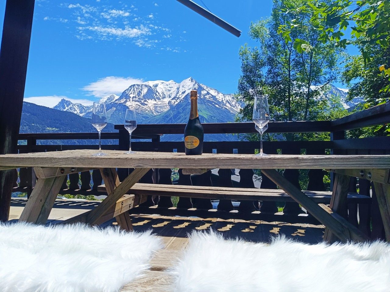 Une table en bois, avec une bouteille et des verres, placée devant un paysage de montagne.
