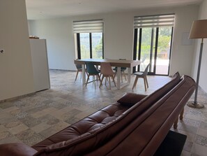 A modern living room with a brown leather sofa, a dining table with chairs, and a large window with blinds. - New, cosy and adult only app in a beautiful village with a lake view (Béznar)