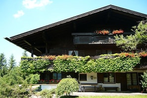 Ein zweistöckiges Holzhaus mit Balkon, umgeben von Grünpflanzen und Bäumen.