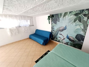 Quarto com sofá azul, cama verde, janela com cortinas leves e parede com papel de parede tropical.