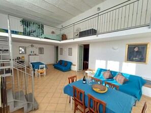 Ein geräumiger Wohnbereich mit Esstisch für zwei Personen, einem blauen Sofa und einer Treppe, die zu einer oberen Ebene führt.