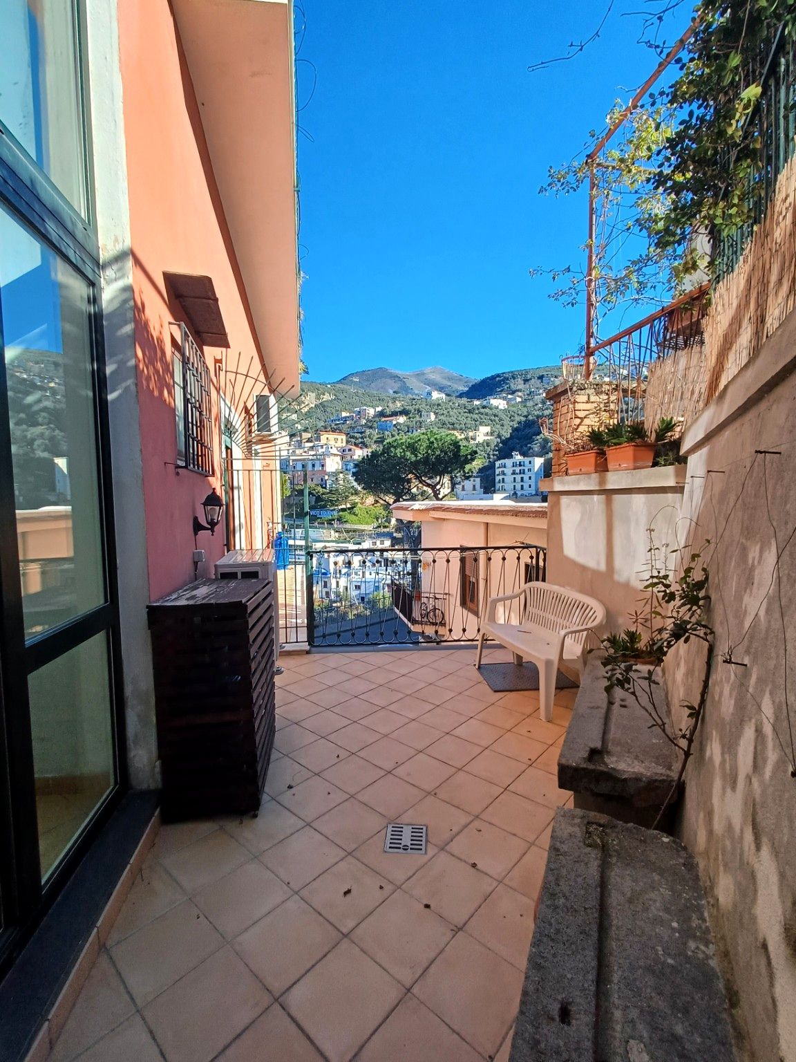 A balcony with a view of a hillside town, a white plastic chair, and a small planter with plants.
