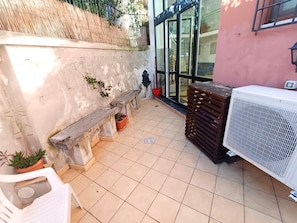 Um pátio com piso de azulejos, um banco, um vaso de planta, um balde vermelho e um aparelho de ar condicionado branco.