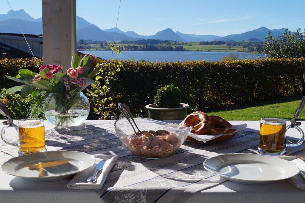 A table set outdoors with a view of mountains, a lake, and a hedge.