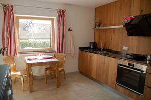 A compact kitchen with a dining table, chairs, a stove, and a window with curtains. - Holiday apartment with 1 bedroom (Hopfen am See)