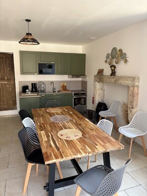 A dining area with a wooden table, black and white chairs, a kitchen in the background, and a hanging light fixture.