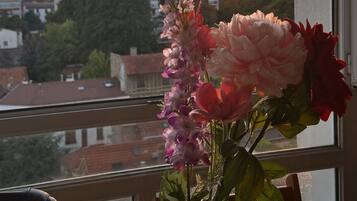 Un vase rempli de fleurs est posé sur une table près d’une fenêtre donnant sur un paysage urbain au coucher du soleil.