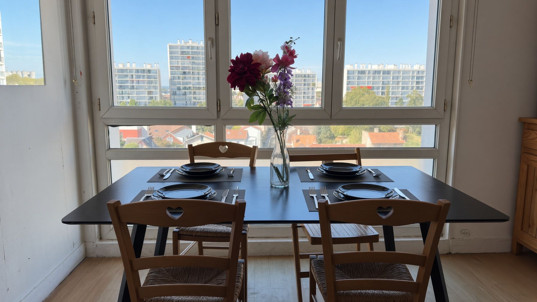 Une table à manger dressée pour deux personnes, avec des chaises en forme de cœur, un vase rempli de fleurs et une vue sur les bâtiments à travers de grandes fenêtres.