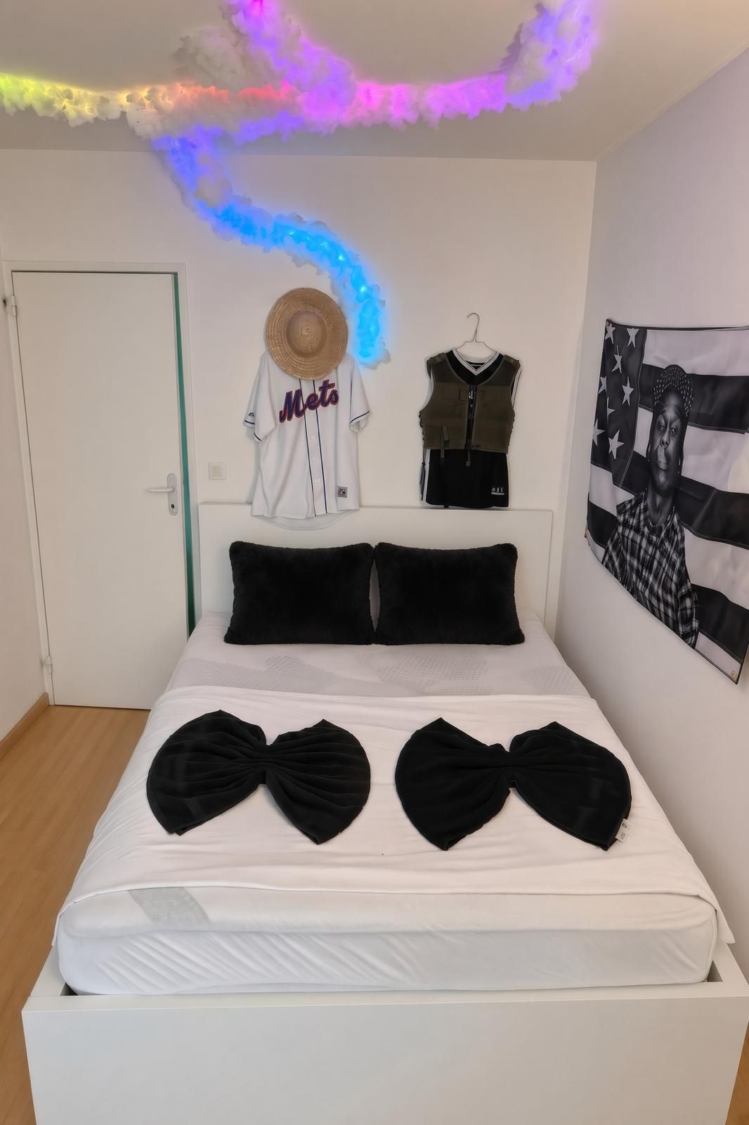 Un letto con cuscini neri e papillon, una maglia bianca, un cappello e un gilet appesi al muro, e una bandiera.