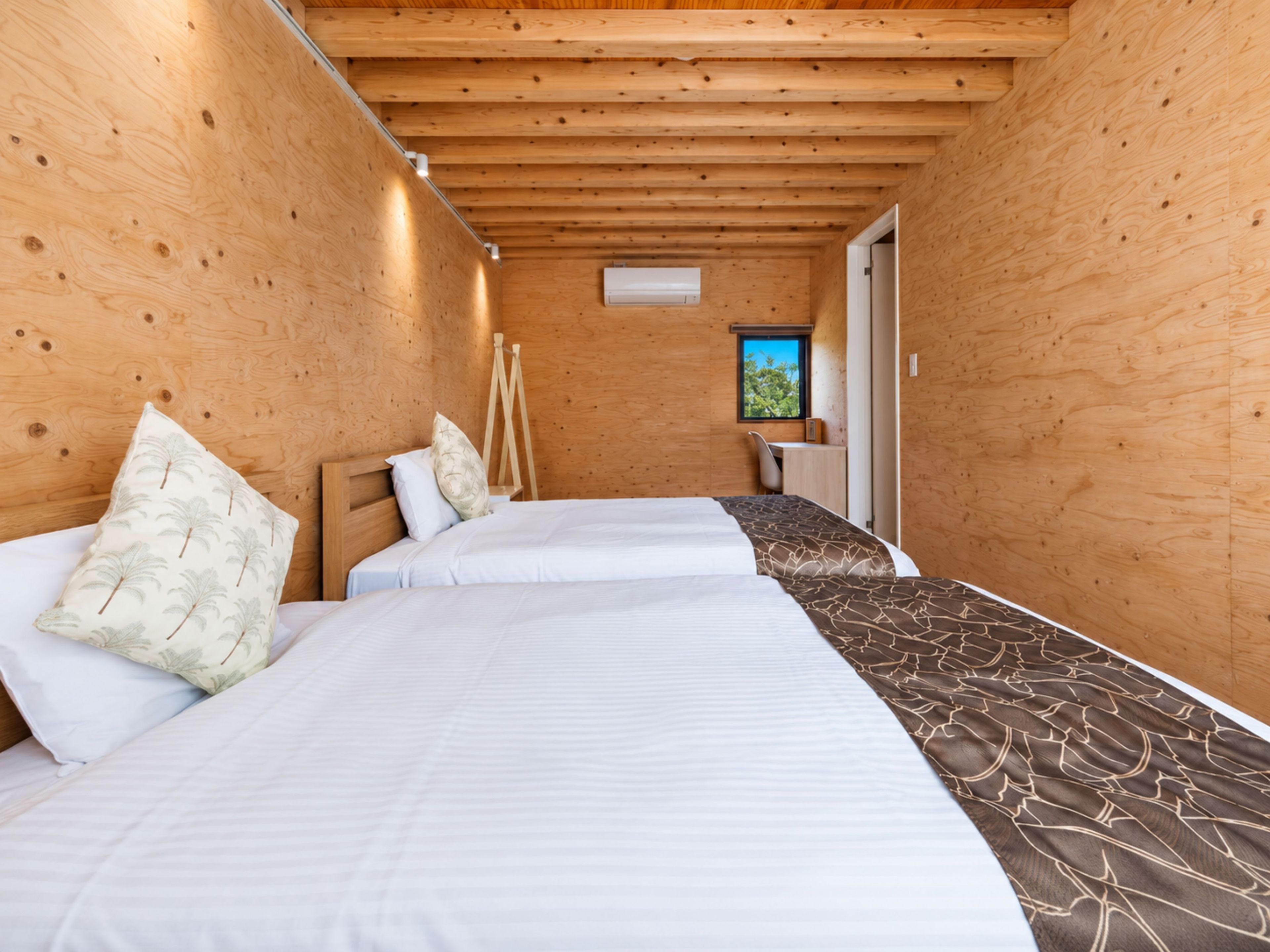 A room with two beds, wooden walls, a small desk, and a window.