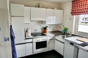 Una cocina con armarios blancos, estufa, horno, lavamanos y una ventana con un cortinero rojo a cuadros.