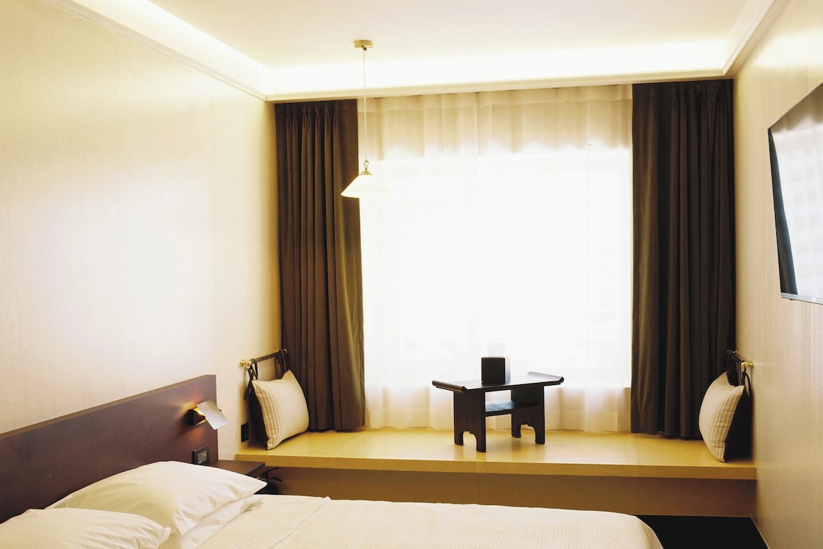 A hotel room with a bed, bedside table, lamp, and a small table with a chair in front of a window with curtains.