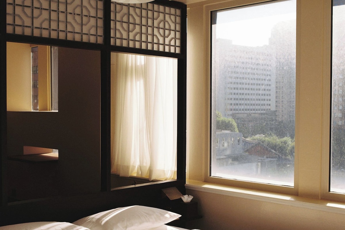 A hotel room with a large window, a bed with white linens, and a hanging light fixture.