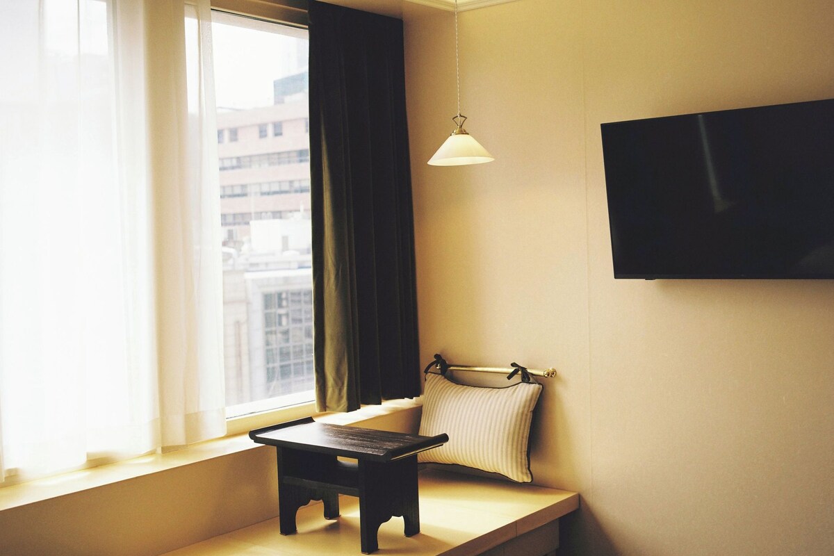 A room with a window, a small table, a hanging light fixture, and a television mounted on the wall.