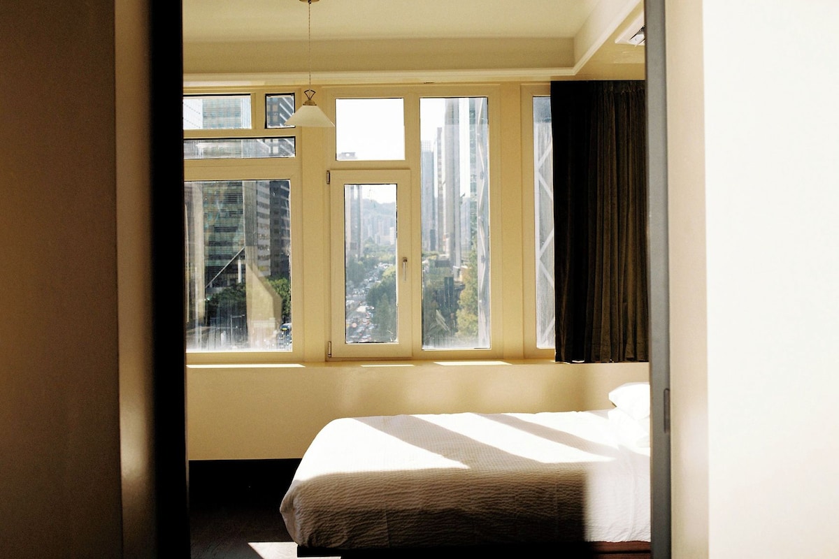 A hotel room with a bed, a window with curtains, and a view of buildings outside.