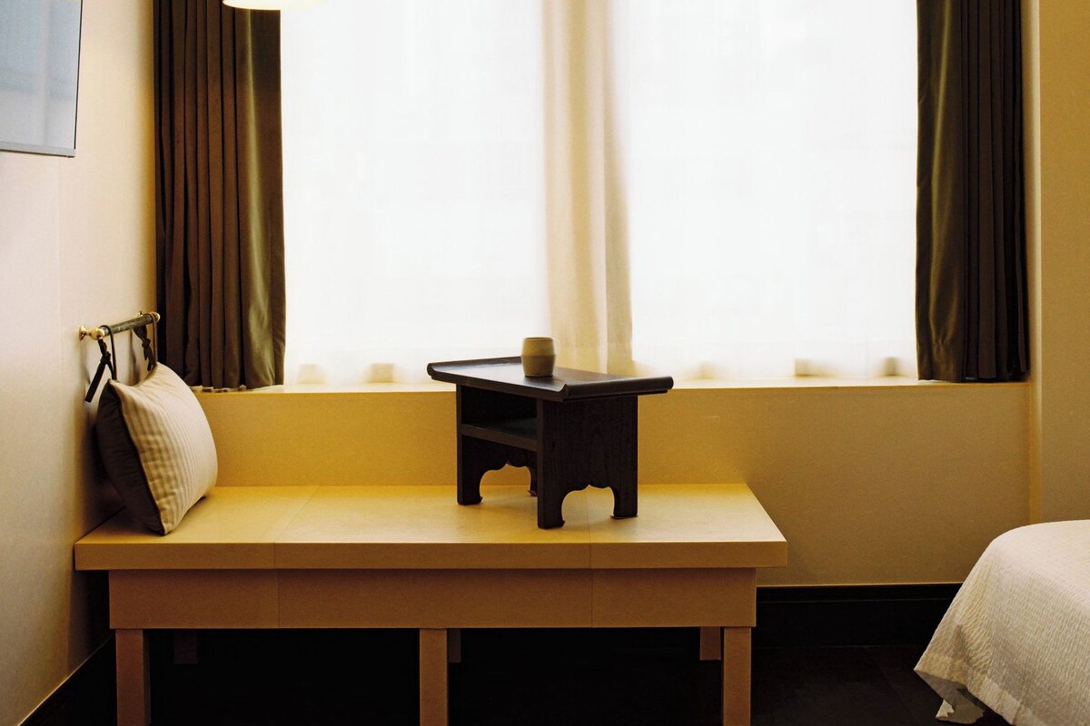 A hotel room with a wooden bench, a small table, a bed, and a window with curtains.