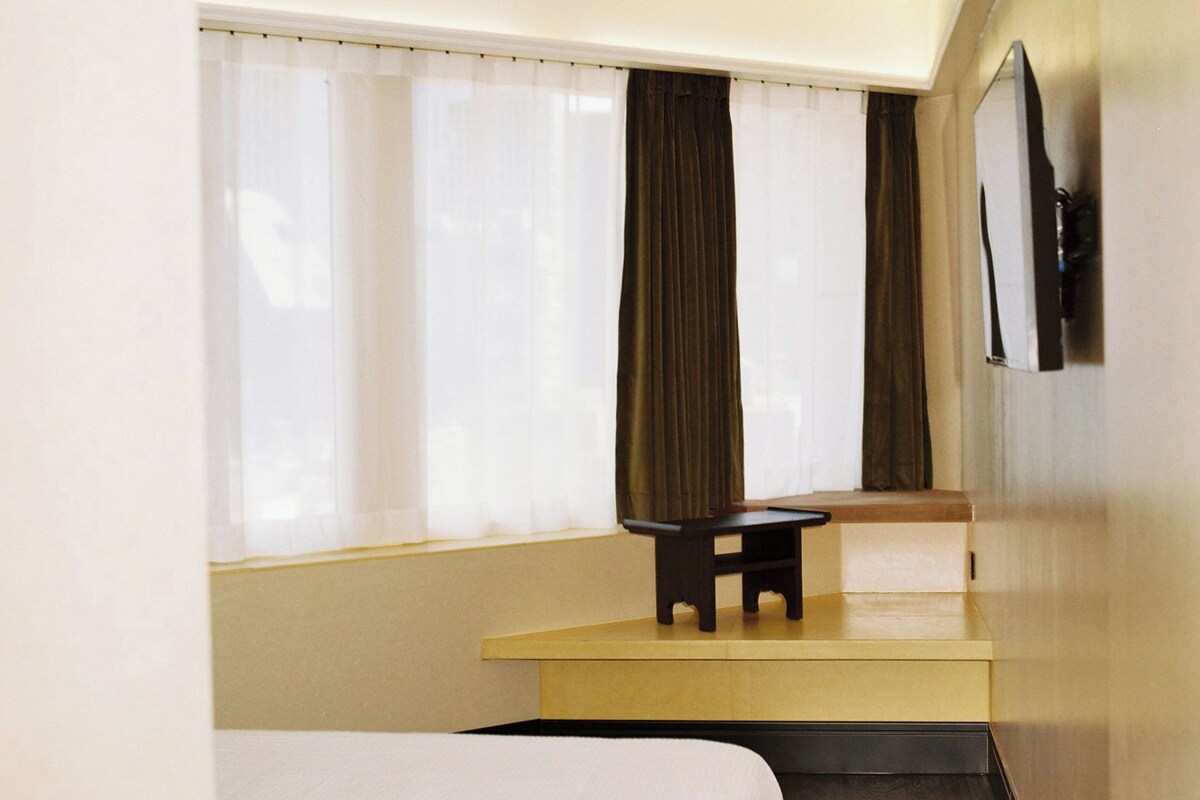 A hotel room with a bed, a small table, a bench, and a window with curtains.
