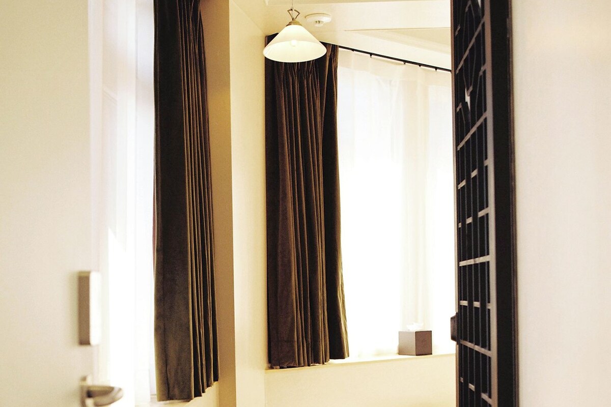 A narrow hallway with a wooden bench, a trash can, a window with curtains, and a hanging light fixture.