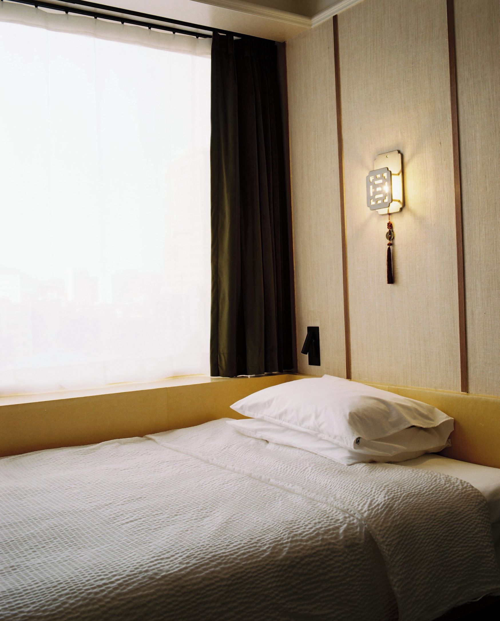 Una habitación de hotel con una cama, una ventana con cortinas, una lámpara de pared y un cabecero de madera.