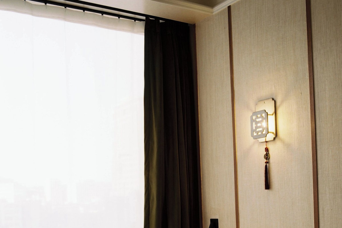 Quarto de hotel com cama, janela com cortinas, luminária fixa na parede e cabeceira de madeira.