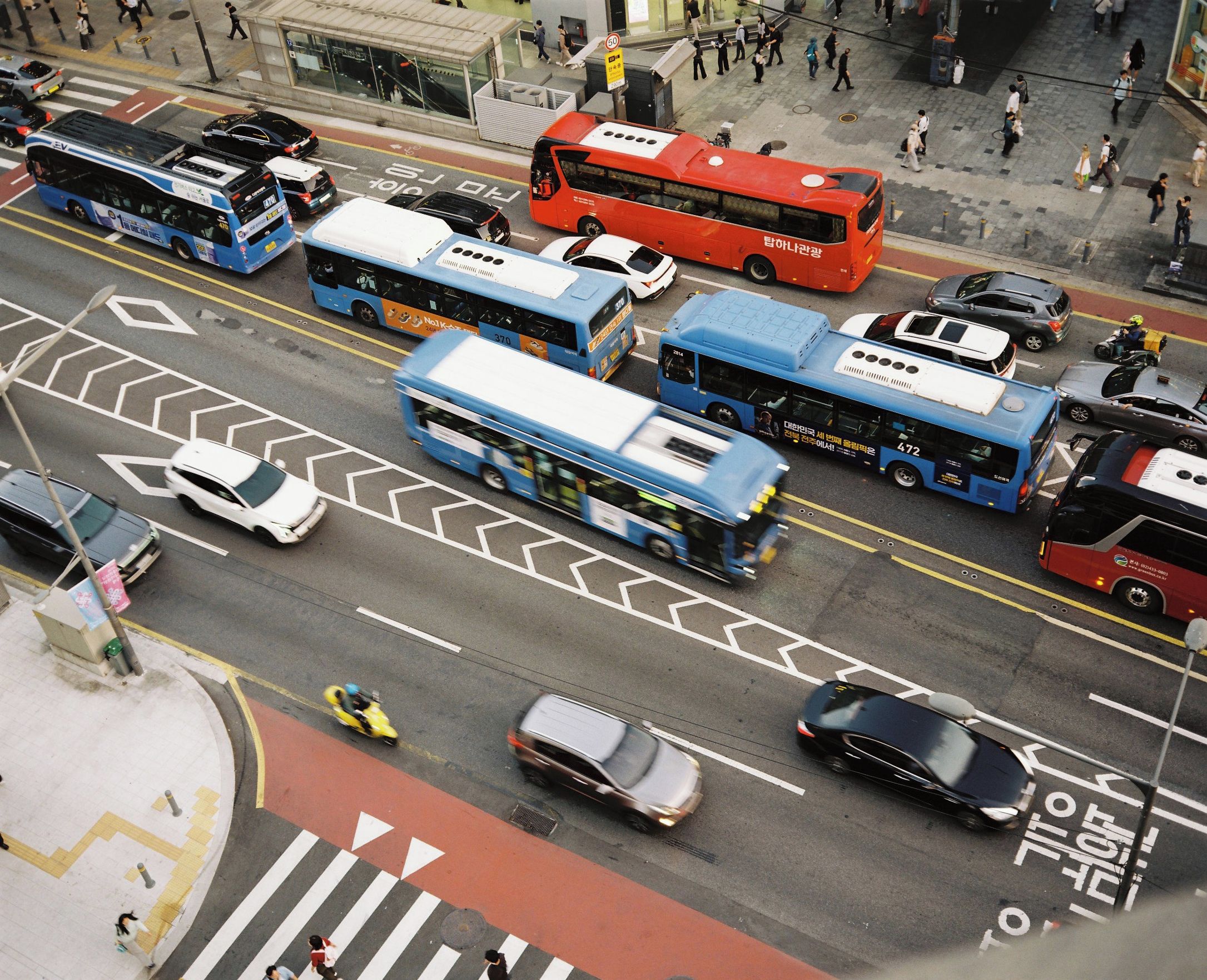 Uma rua movimentada no centro da cidade, com vários ônibus e carros, faixas de pedestres e sinalização viária clara.