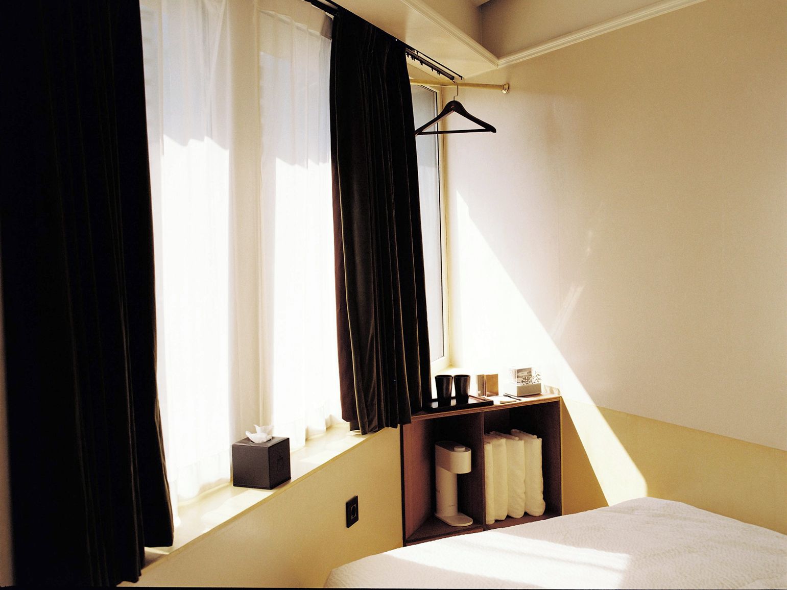 Une chambre d’hôtel avec un lit, une fenêtre avec des rideaux, une table de chevet avec une boîte de mouchoirs et un porte-manteau.