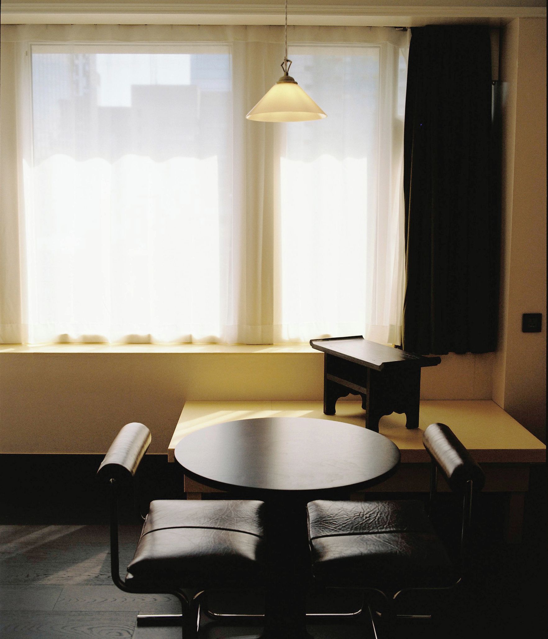 Una habitación con una mesa redonda y dos sillas, un pequeño banco, una lámpara colgante y una ventana con cortinas.