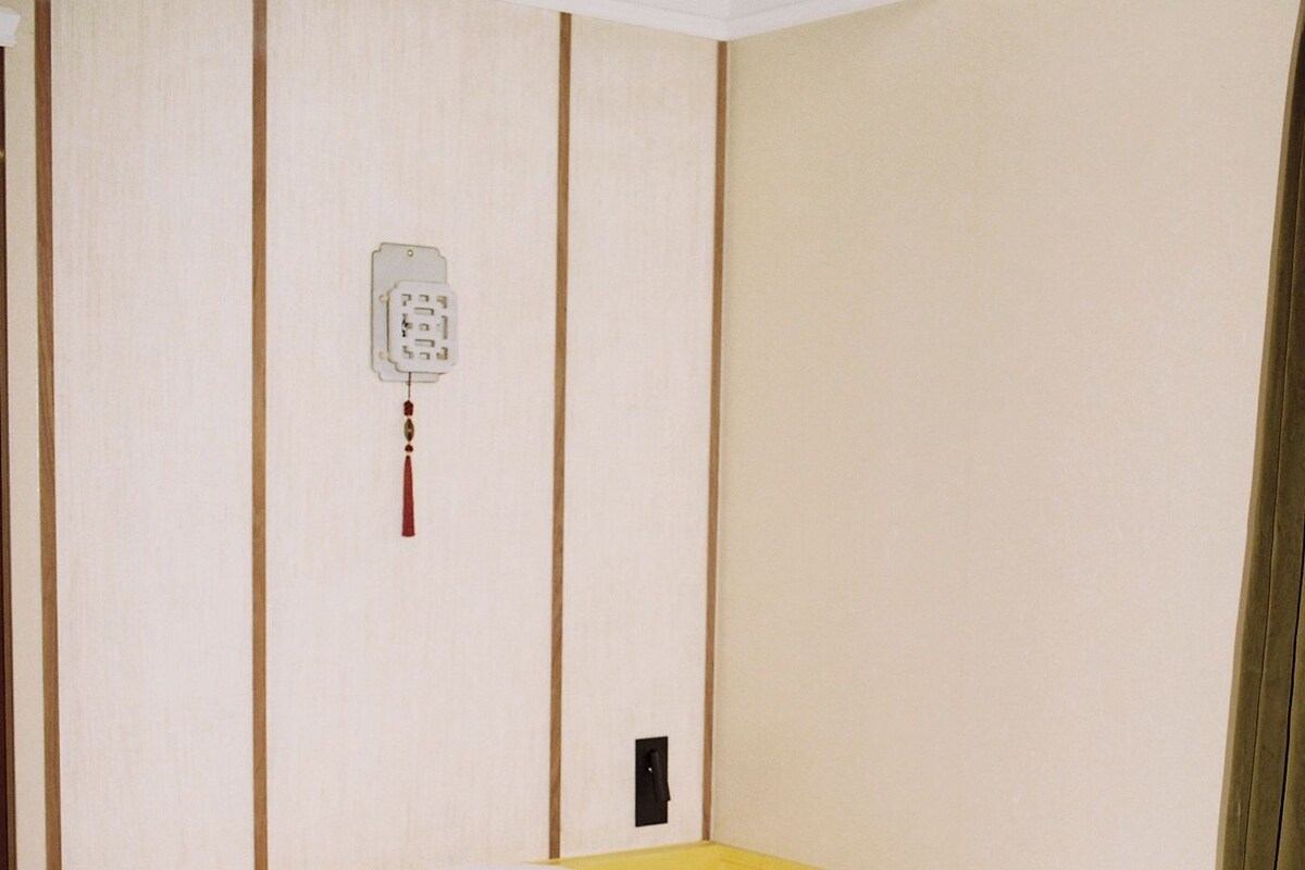 A hotel room with two single beds, a plain wall with a telephone and a light switch, and a curtain on the window.