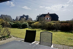 A patio area with a table and chairs, a hedge, and a row of houses in the background. - House Rungholt Holiday Apartment 8 (Wenningstedt-Braderup)