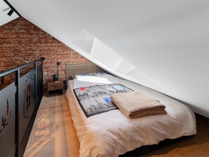 Ein Schlafzimmer mit schrägem Dach, einem Bett mit bedruckter Decke, einem Nachttisch, einer Lampe und einer Ziegelwand.