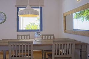 A dining area with a wooden table and chairs, a hanging light fixture, a window with a view of greenery, and a wall-mounted clock.