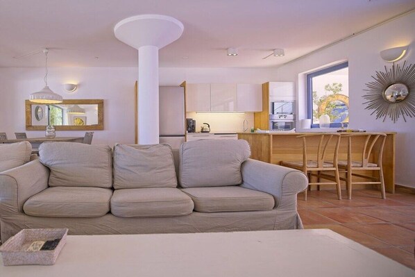 A living room with a beige sofa, a dining area with wooden chairs, and a kitchen with white cabinets.