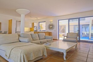 A spacious living area with a sofa, armchair, and coffee table, a kitchen in the background, and a pool visible through the glass doors.