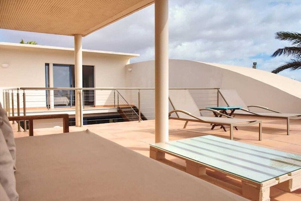 A modern outdoor patio with a glass table, lounge chairs, and a staircase leading to an upper level.