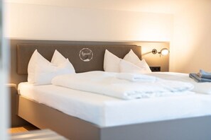 Ein ordentlich gemachtes Bett mit weißen Leinen und Kissen, ein Kopfteil mit einem Logo sowie eine Nachttischlampe.