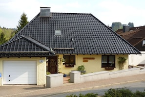A single-story building with a dark tiled roof, a garage door, and a small porch area with potted plants. - Holiday apartment with 2 bedrooms (St. Andreasberg)