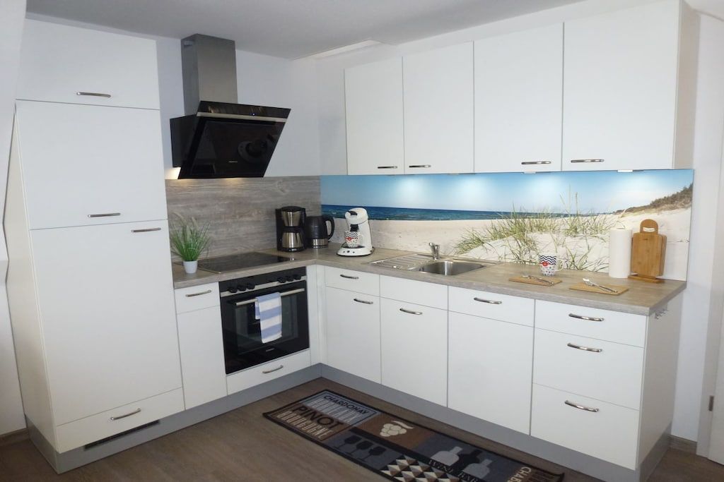 Eine moderne Küche mit weißen Schränken, einer Edelstahl-Spüle, einer Kaffeemaschine und einer Spritzschutzleiste mit Strandmotiv.