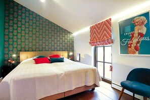Une chambre à coucher avec un lit, des oreillers, une table de chevet, une chaise, une fenêtre donnant sur un paysage agréable et un mur orné d’une affiche.