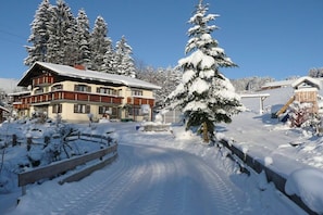 A snow-covered building with a wooden fence, a snow-covered tree, and a clear sky. - Guesthouse Schmid & Reitzner 2 (Immenstadt)