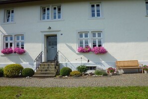 A white building with a front door, windows, and flower boxes. There are steps leading to the door, a bench, and a bird on the roof. - Holiday apartment with 2 bedrooms (Schwirzheim)