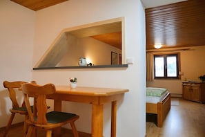 A room with a wooden dining table, chairs, a small potted plant, a bed with a checkered bedspread, and a window with curtains. - Guesthouse Schmid & Reitzner 3 (Immenstadt)