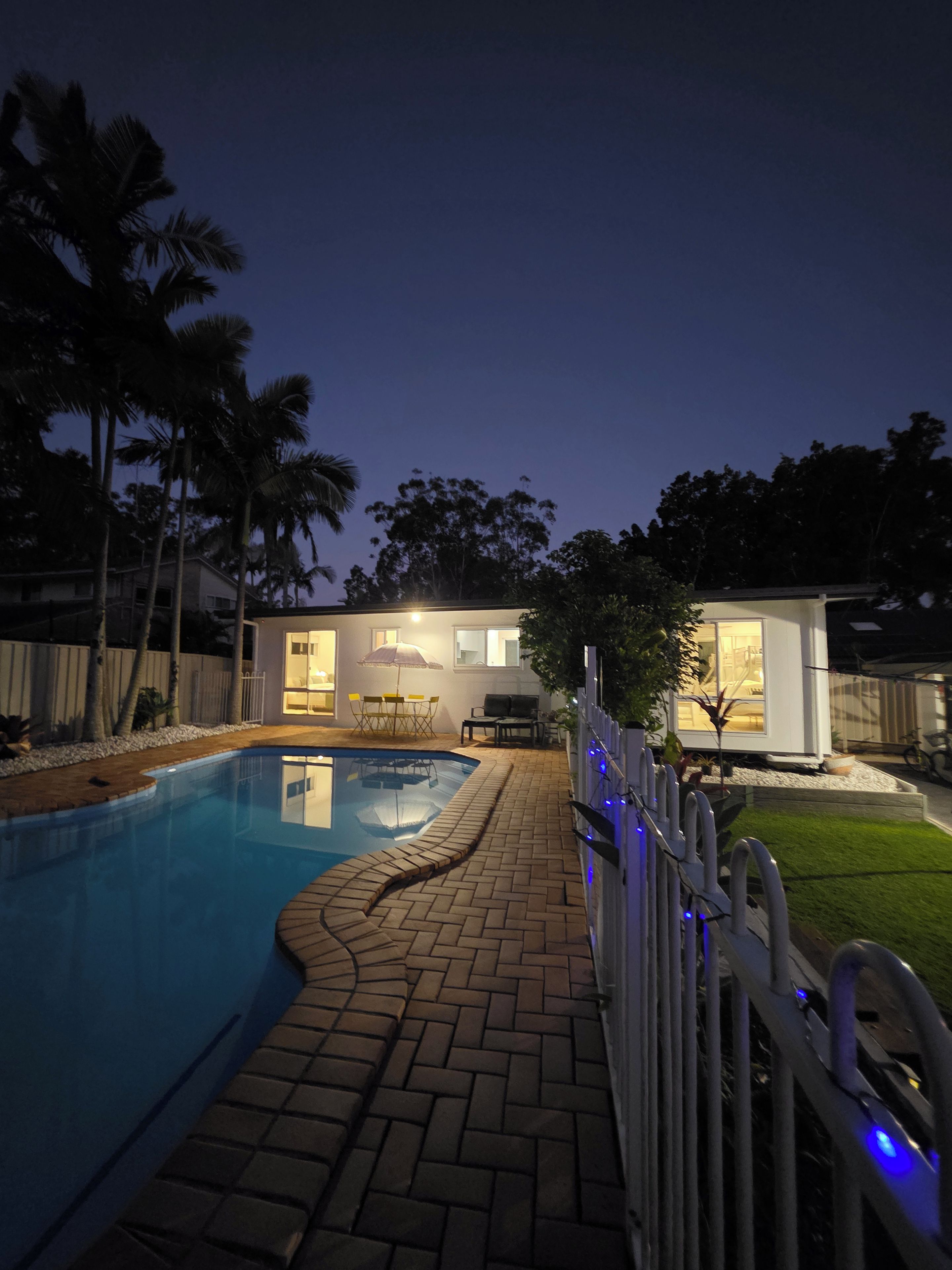 A swimming pool with a brick deck, a fenced area, and a house with a patio and outdoor furniture.
