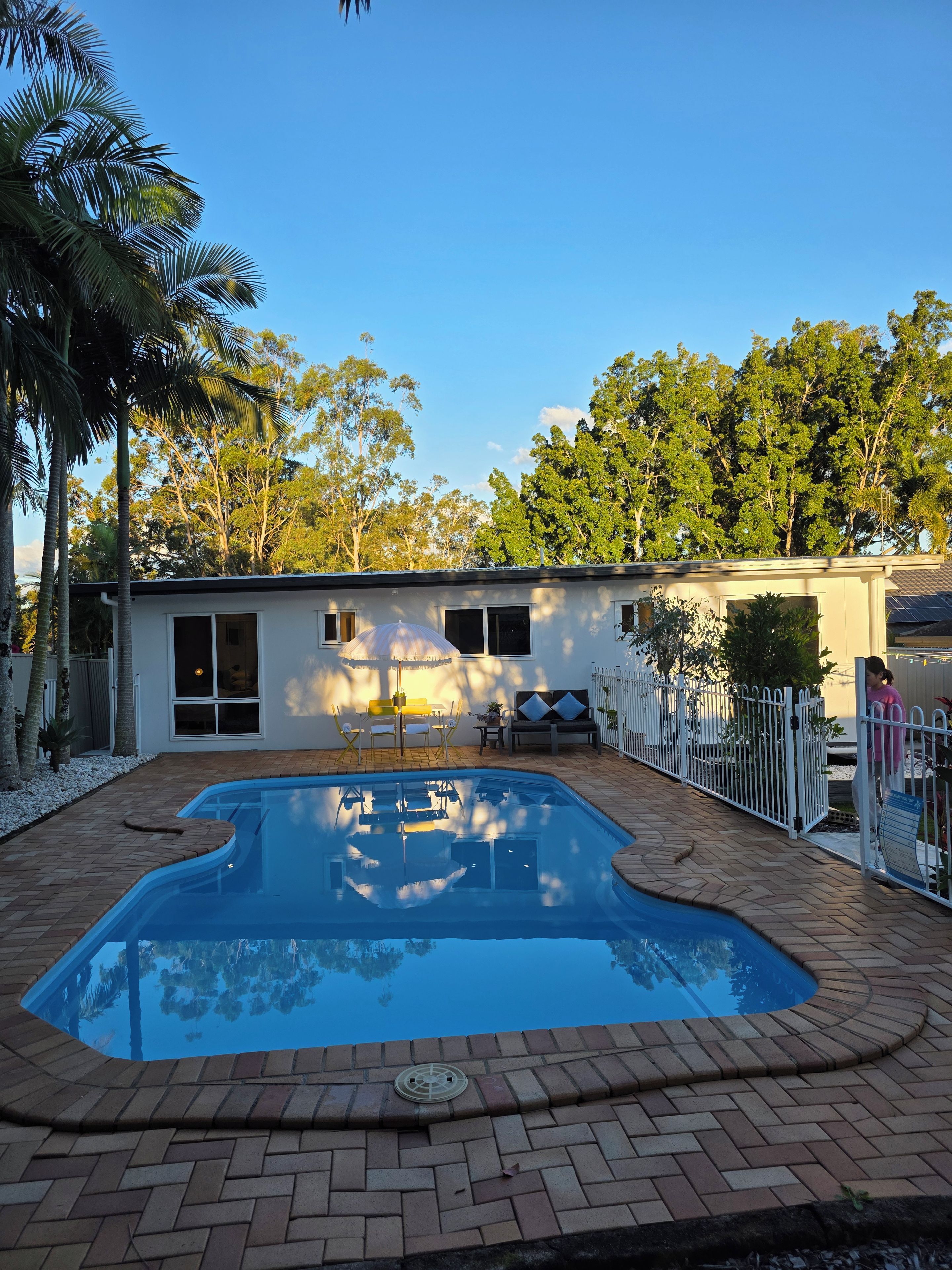 A swimming pool with a brick border, a small building with a window and a door, and a white fence.