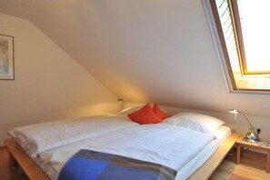 Ein Einzelbett mit weißer Bettwäsche und einem roten Kopfkissen in einem Zimmer mit schrägem Dach und Dachfenster.