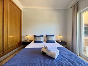 Ein Hotelzimmer mit einem großen Bett, zwei Nachttischen mit Lampen, einem Holzschrank und Blick ins Freie durch ein Fenster.