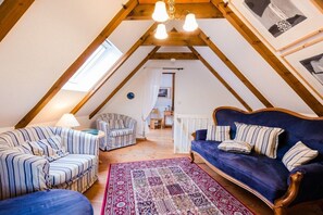 Ein Loft-Zimmer mit schrägem Dach, einem blauen Sofa, einem gestreiften Sessel, einem gemusterten Teppich und einer Holzbalkenkonstruktion an der Decke.