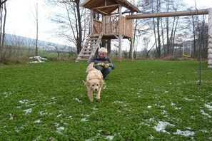 A person sitting on the grass with a dog in a park with a wooden play structure. - On the Freimuth Maria farm (Neukirchen b. Hl. Blut)
